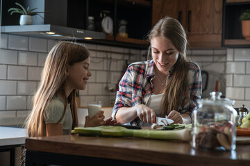 Mother prepares ingredients for making the meal while her daughter stands by the kitchen table and drinks milk.	
