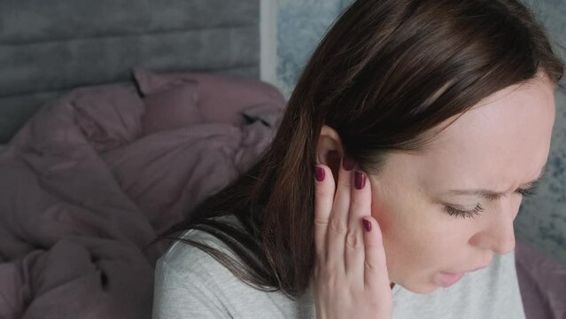 Woman suffering from earache or tinnitus sitting on a bed at home. Caucasian female with ear pain or ear ache. Close up. Tinnitus, otitis, meningitis concept