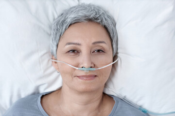 High angle view of senior woman in oxygen tube in her nose looking at camera while lying on bed in...