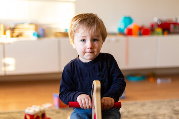 Selective focus view of fair toddler boy sitting on rocking horse in family room in soft morning light