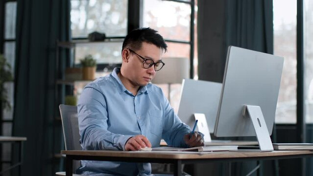 Serious middle aged asian man in eyeglasses working at office, comparing information on computer screen and documents