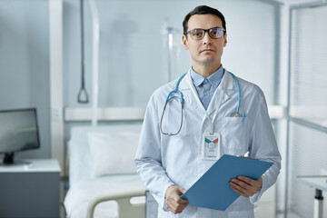 Portrait of young doctor in white coat with medical card looking at camera while standing in ward...