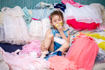 Little girl chooses clothes while sitting on the couch. Different outfits to choose from in the dresser. Shopaholic bought herself a lot of things.