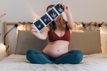 white, blonde pregnant woman sitting cross-legged on the bed shows pictures of the fetus taken with...