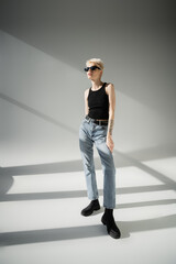 Fototapeta premium full length of tattooed model in black sunglasses and jeans posing on grey background.