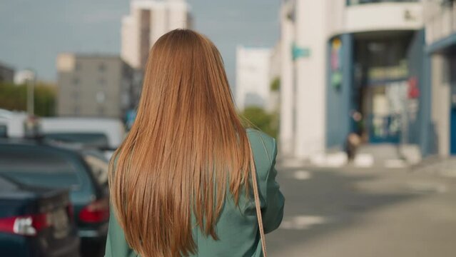 Woman with long loose red hair talks on phone walking along car parking in big city. Businesswoman conducts business remotely against blurry buildings backside view