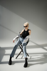 full length of stylish blonde woman in sunglasses and trendy outfit sitting on folding chair on grey.