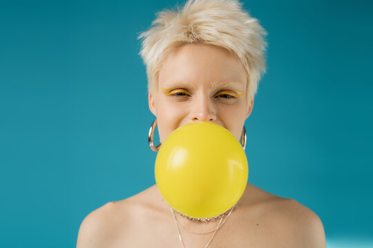 blonde albino woman with bare shoulders blowing bubble gum on blue background.