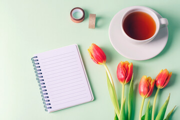 Spiral notepad, a cup of tea and red tulip flowers on light green background. Top view, flat lay,...