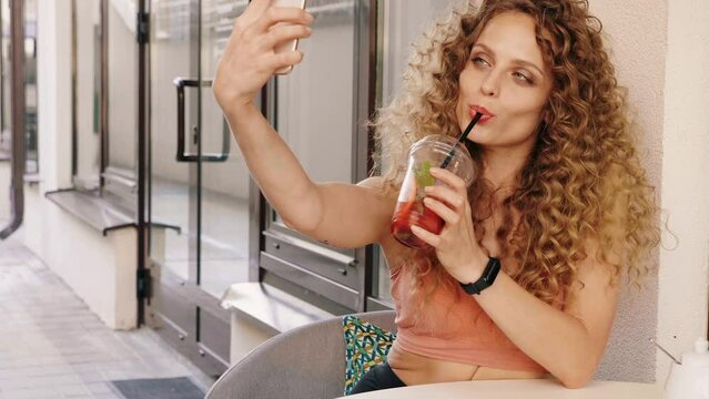 Young beautiful smiling hipster woman with afro curls hairstyle. Woman in summer trendy clothes sitting in veranda cafe. Model drinking fresh cocktail smoothie drink in plastic cup with straw