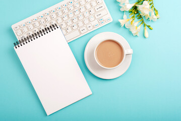 Obraz na płótnie Canvas Computer keyboard, blank notepad and coffee cup on blue table with freesia flowers. Top view, flat lay, mockup