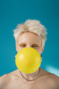 blonde albino woman with yellow eye liner blowing bubble gum on blue background.