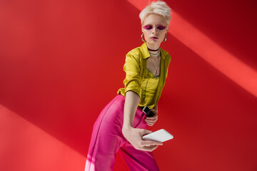 albino woman in trendy sunglasses taking selfie on smartphone on carmine pink background.