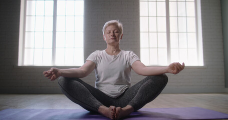 Fototapeta premium Mature caucasian woman doing yoga in studio. Senior woman relaxing in lotus position on floor, keeping a healthy lifestyle - wellness concept 
