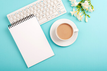 Computer keyboard, blank notepad and coffee cup on blue table with freesia flowers. Top view, flat lay, mockup