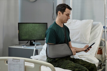 Soldier with injured arm in bandage reading message on smartphone while sitting on bed in hospital...