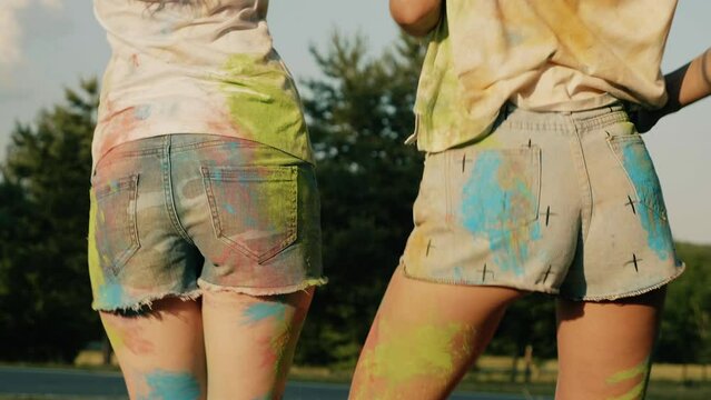 Back view of two girls making party at Holi colours festival in summer time.Young women friends having fun after music event at sunset. Cheerful models smeared with coloured paints in the park