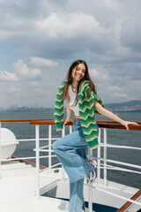 Obraz premium Positive young woman in knitted sweater and jeans standing near railing of yacht in Turkey.