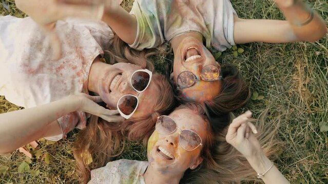 Three happy beautiful female making party at Holi colours festival in summer time. Young  smiling women friends having fun after music event at sunset. Models lying on the grass in sunglasses