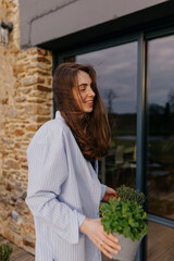 Beautiful young brunette european woman is holding fresh plants on summer terrace and smiling. Model wears shirt and jeans. Lifestyle concept sincere emotions