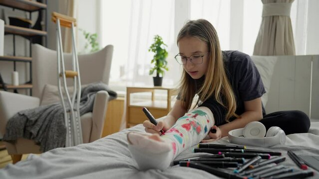 Teenager girl paints on cast leg with felt-tip pens. Young woman draws drawing on cast after injury. Illness, recovery period at home with crutches. Bone fracture. Positive attitude towards situation.