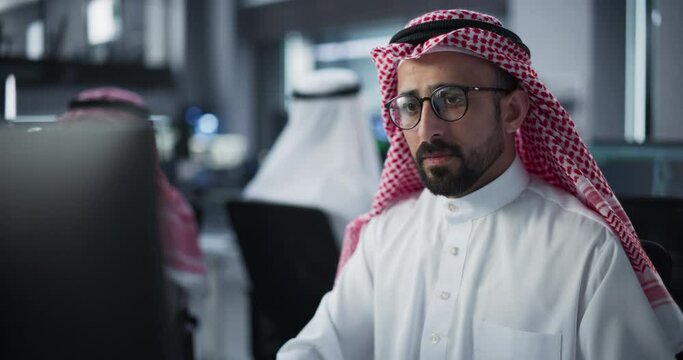 Portrait of a Smart Young Arab Specialist Working in a Technological Startup Help Desk Department, Using Computer in a Diverse Office. Male Wearing Traditional Middle Eastern Clothes