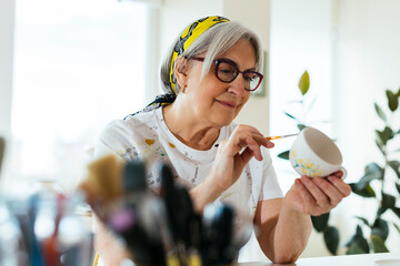 Smiling woman wearing eyeglasses painting ceramic cup in workshop