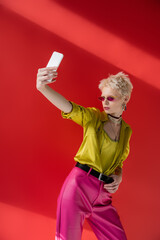 tattooed albino woman in stylish sunglasses taking selfie on smartphone on carmine pink background.