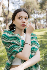 Naklejka premium Portrait of young woman with knitted sweater on shoulders holding daisy in park.