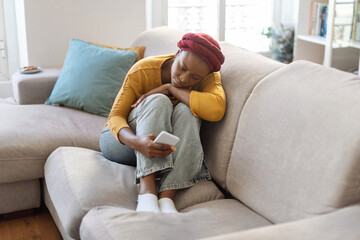 Obraz premium Upset black woman sitting on sofa, using phone, home interior