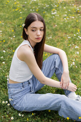 Young woman in top and jeans sitting on lawn with daisy flowers in park.