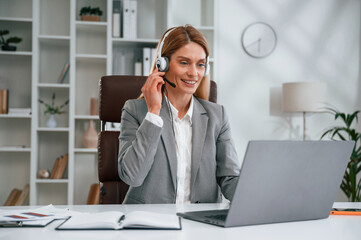 In headphones, by laptop, call center. Woman in business formal clothes is working in office