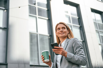 Fototapeta premium With cup of drink and phone. Woman in formal clothes is outdoors near the business building