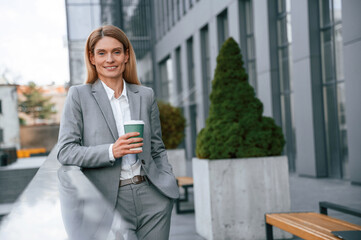 Fototapeta premium Smiling, holding cup of drink. Woman in formal clothes is outdoors near the business building