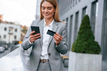 Fototapeta premium Holding credit card and smartphone. Woman in formal clothes is outdoors near the business building