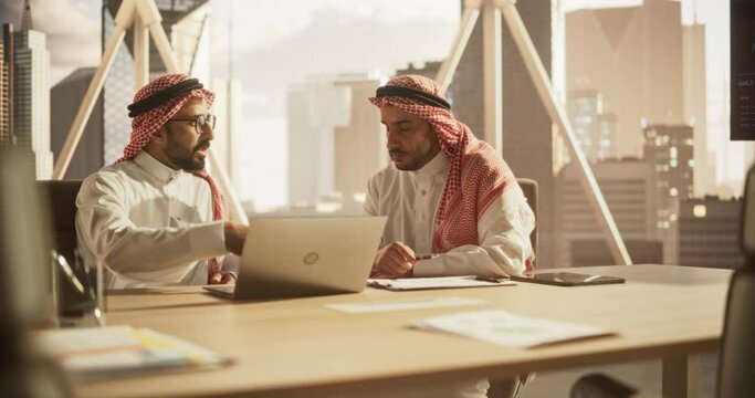 Arab Financial Analyst Having a Meeting to Discuss a Project with a Team Leader. Middle Eastern Colleagues Using Laptop Computer, Working in Modern Office, Wearing Traditional Thobe and Kaffiyeh