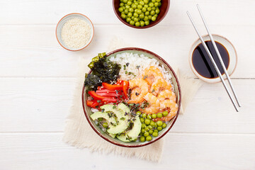 A poke bowl with shrimp, rice, avocado, pepper, green peas, nori seaweed, sesame seeds on a white wooden background. Hawaiian diet dish with seafood. Top view.