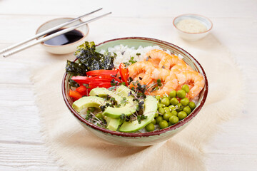 A poke bowl with shrimp, rice, avocado, pepper, green peas, nori and micro greens on a white wooden background. Hawaiian diet dish with seafood. Close-up