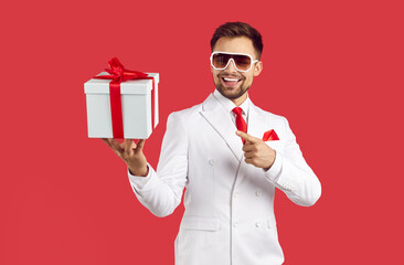 Cheerful stylish young man in suit points to gift box in his hands isolated on red background....