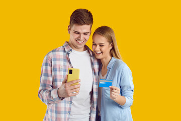 Young positive Caucasian man and woman are engaged in joint Internet shopping by paying for purchase in apps of mobile phone with credit card dressed in casual clothes stands in yellow studio