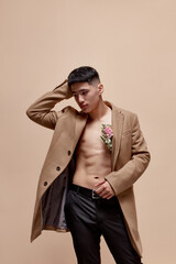 Fototapeta na wymiar Portrait of handsome young asian guy posing in beige coat on shirtless body with flowers against light brown studio background. Fashion, style, body aesthetics, beauty, men's health, emotions concept