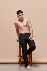 Portrait of young handsome guy with muscular, relief body posing shirtless in black trousers, leaning on chair against light brown studio background. Fashion, body aesthetics, beauty, emotions concept