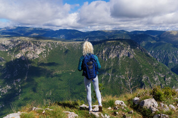 Naklejka na ściany i meble An unrecognisable female tourist with a backpack on her back looks at the mountains from a cliff