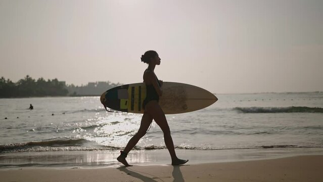 African american woman walking with surfboard on ocean beach. Black female surfer going with surf board. Pretty multiethnic girl goes on surfing session at sunrise. Slow motion, side view.