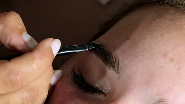 Hand of professional master putting gel with tiny brush on eyebrow of female client. Young woman lies with closed eyes in beauty salon closeup