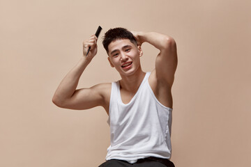 Getting ready. Portrait of handsome smiling young guy with muscular body, brushing hair against light brown studio background. Concept of male beauty, skincare, cosmetology, men's health