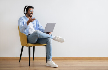 Happy young middle eastern guy in casual and glasses, wireless headphones has video call on laptop
