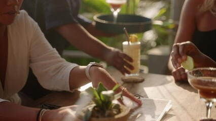 Woman waiter with tray serves cocktails to multiracial women in sea view tropical restaurant. Female hand takes glasses with alcohol drinks from puts them onto table of multiethnic smiling females