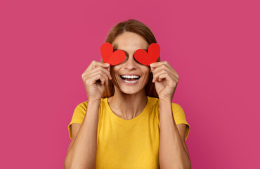 Laughing funny middle aged european blonde lady puts hearts to her eyes, like glasses, has fun