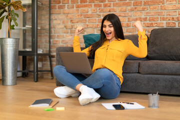 Excited teen spanish lady shaking fists, getting excellent mark on test, sitting on floor and using laptop computer